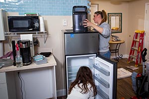  Design team member sets up equipment on top of a refrigerator in the kitchenette, while the designer kneels down to place shelves in the refrigerator